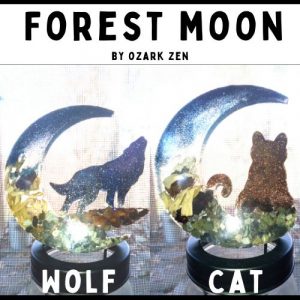 A photo of a two resin wall art pieces in the shape of a crecent moon, one with a wolf and one with a cat, the wall art features natural elements from the forest including hemlock bark, heirloom sword fern fronds, salt water stones, and biodegradable glitter and inks. The title is Forest Moon, this Branson art piece is by Ozark Zen in Branson Missouri.